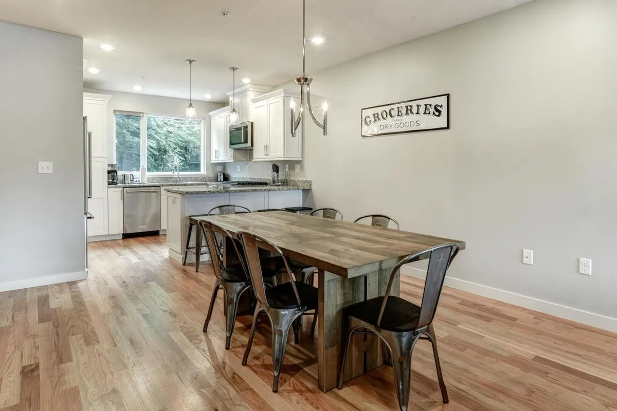 A welcoming dining area with a large table and kitchen island, illuminated by daylight pouring through a window that offers a partial view of the outdoors. The layout suggests a space ideal for casual gatherings, shared meals, and lively conversations before or after a night out on the town.