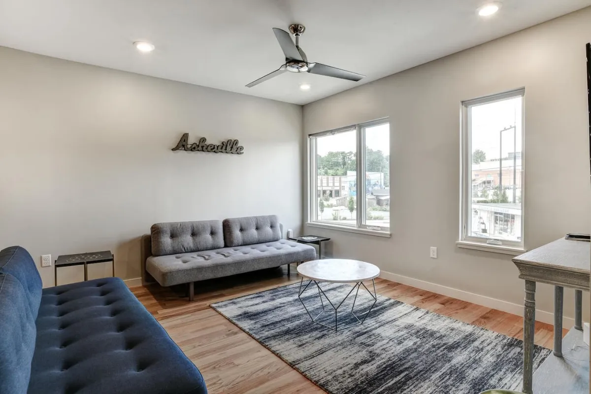 A bright and comfortable living area featuring a cozy sofa, a modern coffee table, large windows that fill the space with natural light, and a decorative sign that adds a welcoming touch. The arrangement encourages socializing and provides a stylish, laid-back atmosphere for guests to gather.