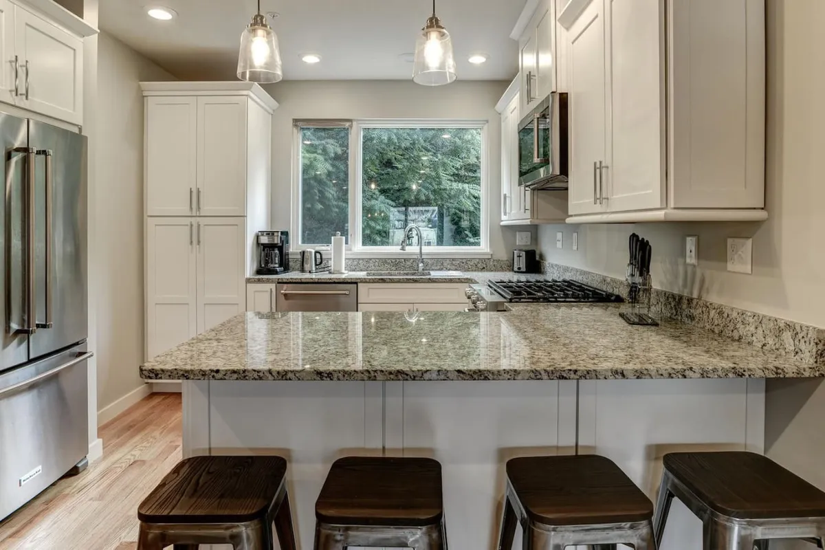 A bright and welcoming kitchen featuring a granite countertop and central island, flooded with natural light from a large window overlooking greenery. The open space suggests ease of movement and a comfortable gathering spot.