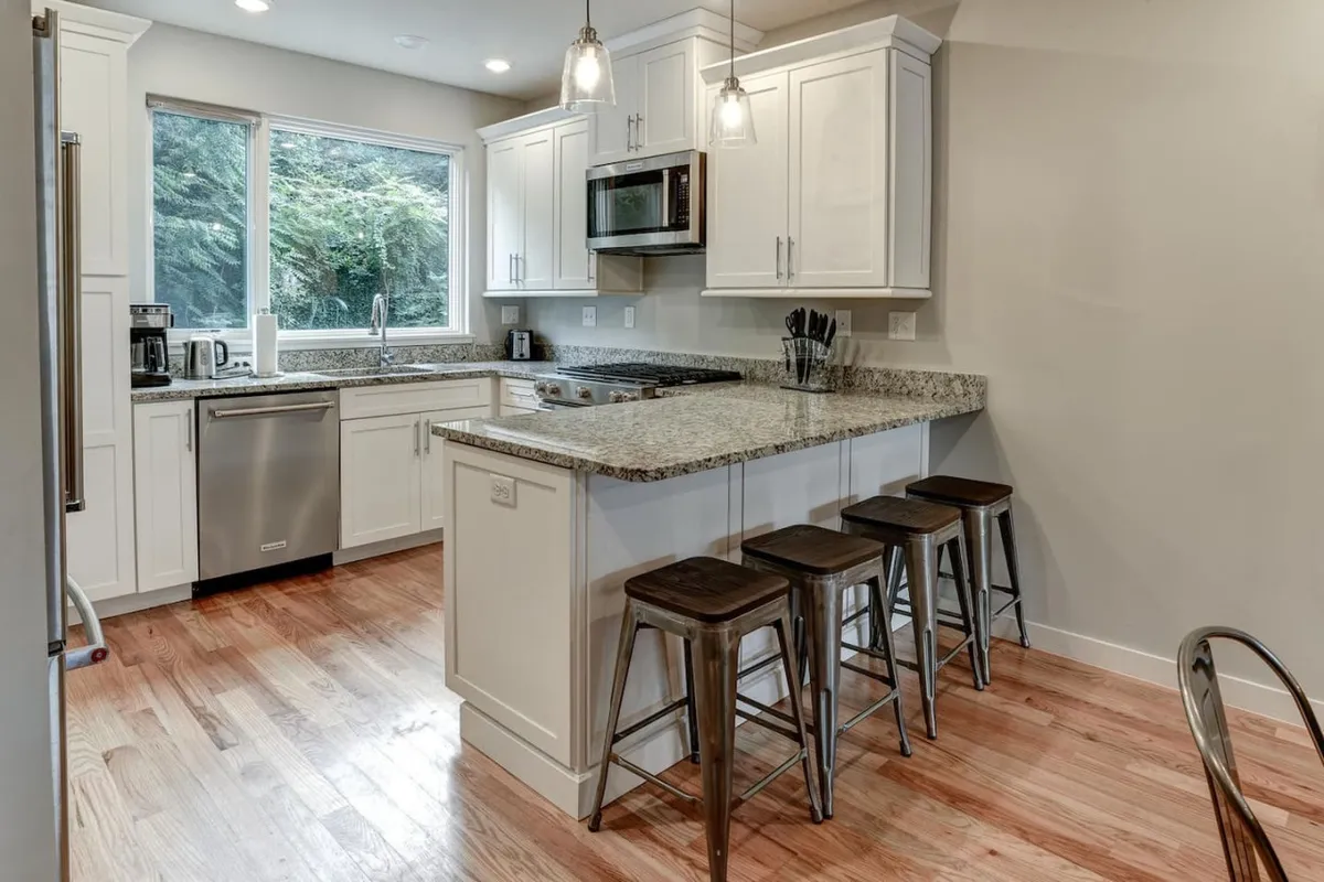 This image highlights a modern, open kitchen with a spacious island and stools, stainless steel appliances, and a large window that brings in natural light and frames greenery outside. The view shows the kitchen as a central, functional space, ideal for cooking, entertaining, and casual dining.