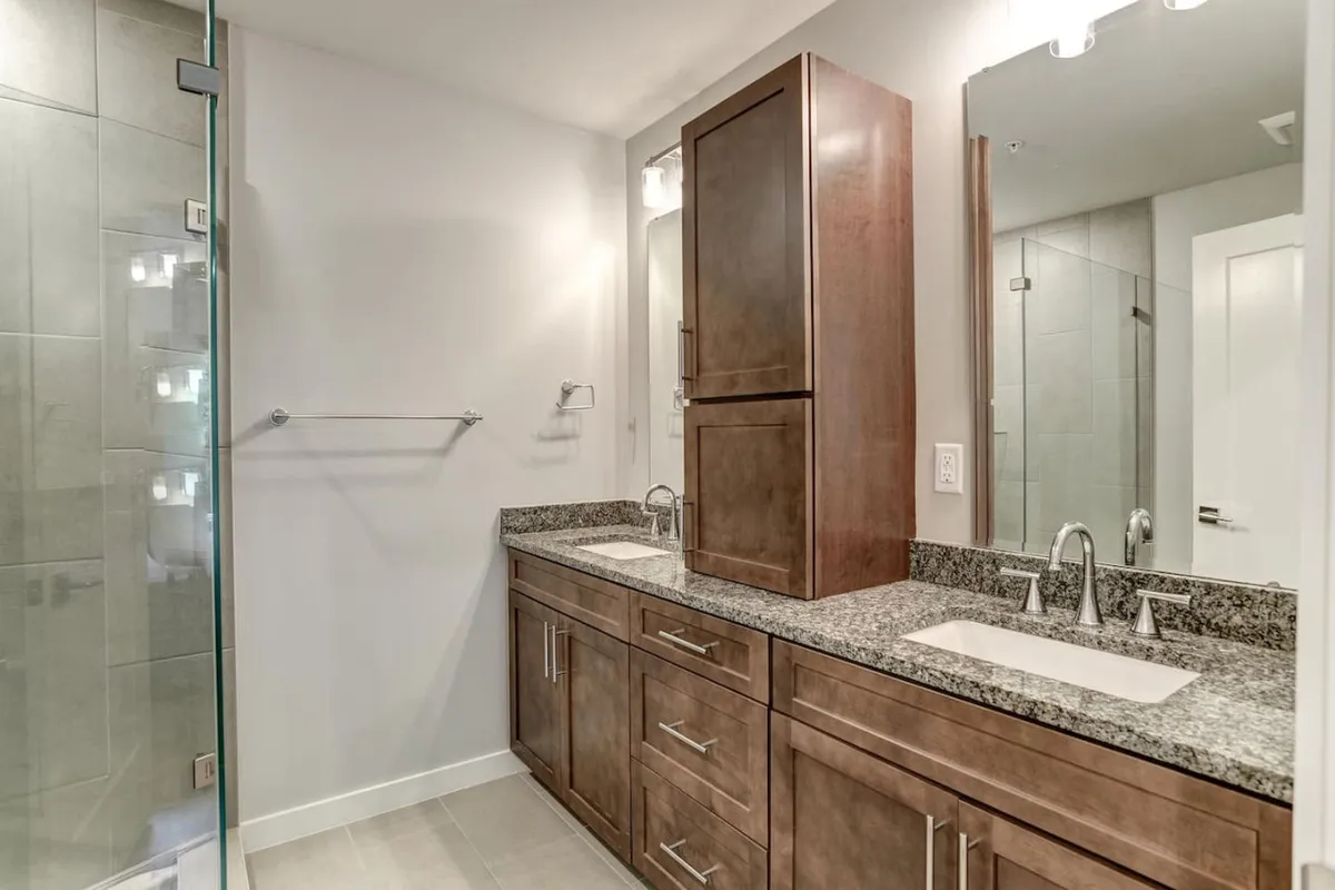 A modern bathroom featuring a double sink vanity, granite countertop, and a glass shower enclosure. The space is well-lit and organized, evoking a spa-like, calm atmosphere ideal for self-care routines.