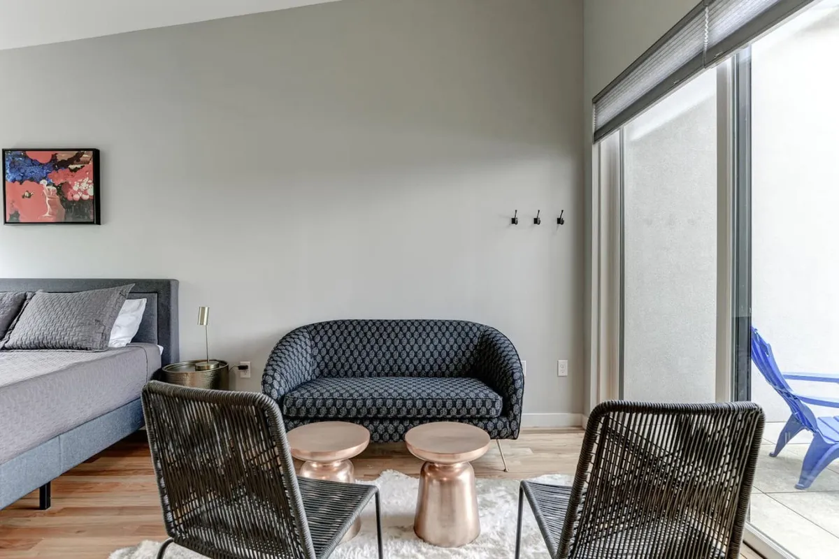 A stylish living area featuring a bed with decorative pillows, a blue sofa, and coffee tables positioned beside sliding glass doors. Natural light floods the space, and the doors suggest access to a patio or the outdoors, reinforcing the link to rooftop evenings or city explorations.