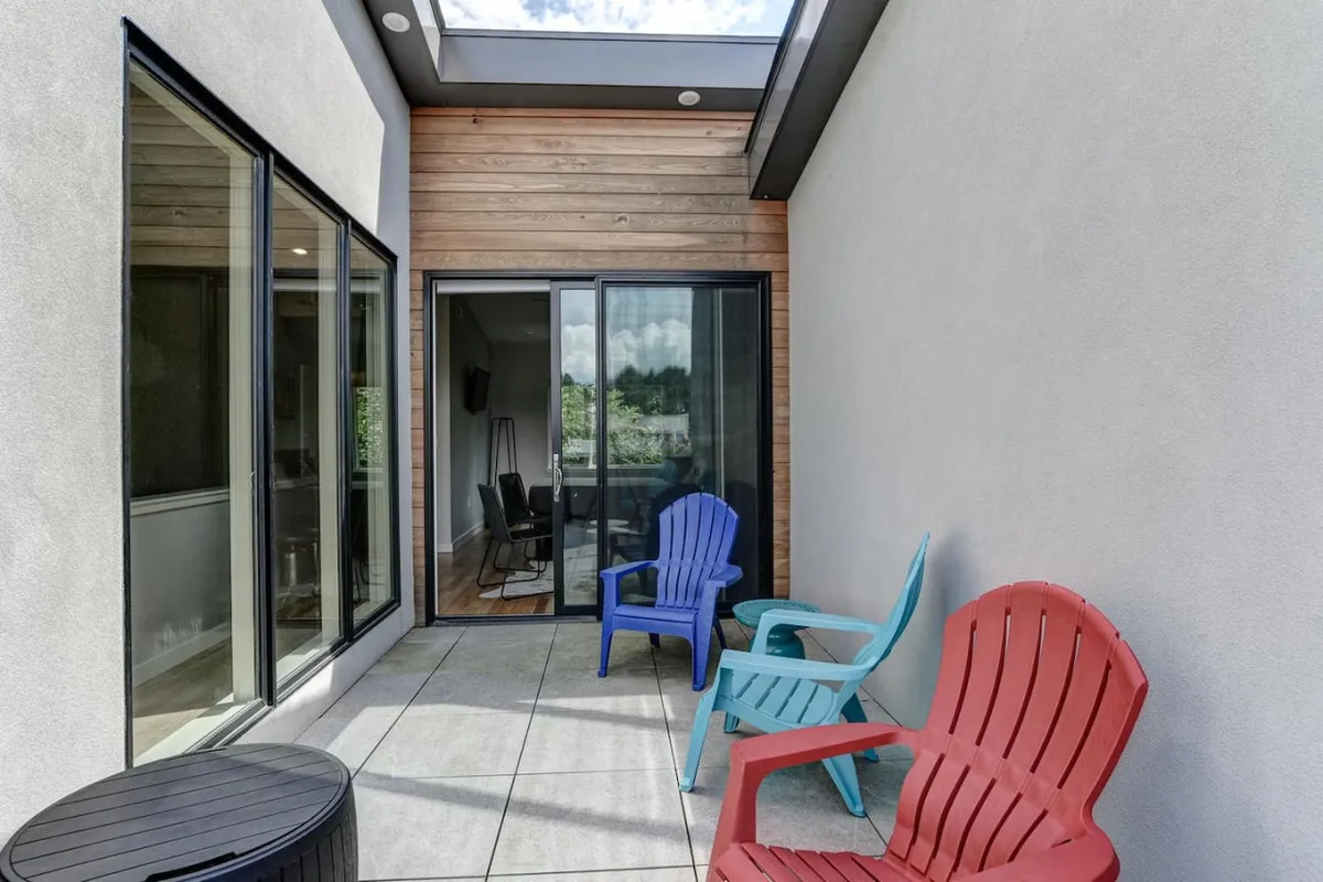 A rooftop patio scene featuring colorful chairs arranged around a small table, set against a wooden wall and accessed via a sliding glass door. The space is bathed in sunlight, suggesting a perfect spot for relaxing or gathering with friends. The open sky overhead hints at wide views, ideal for sunrise coffee, sunset cocktails, or stargazing.