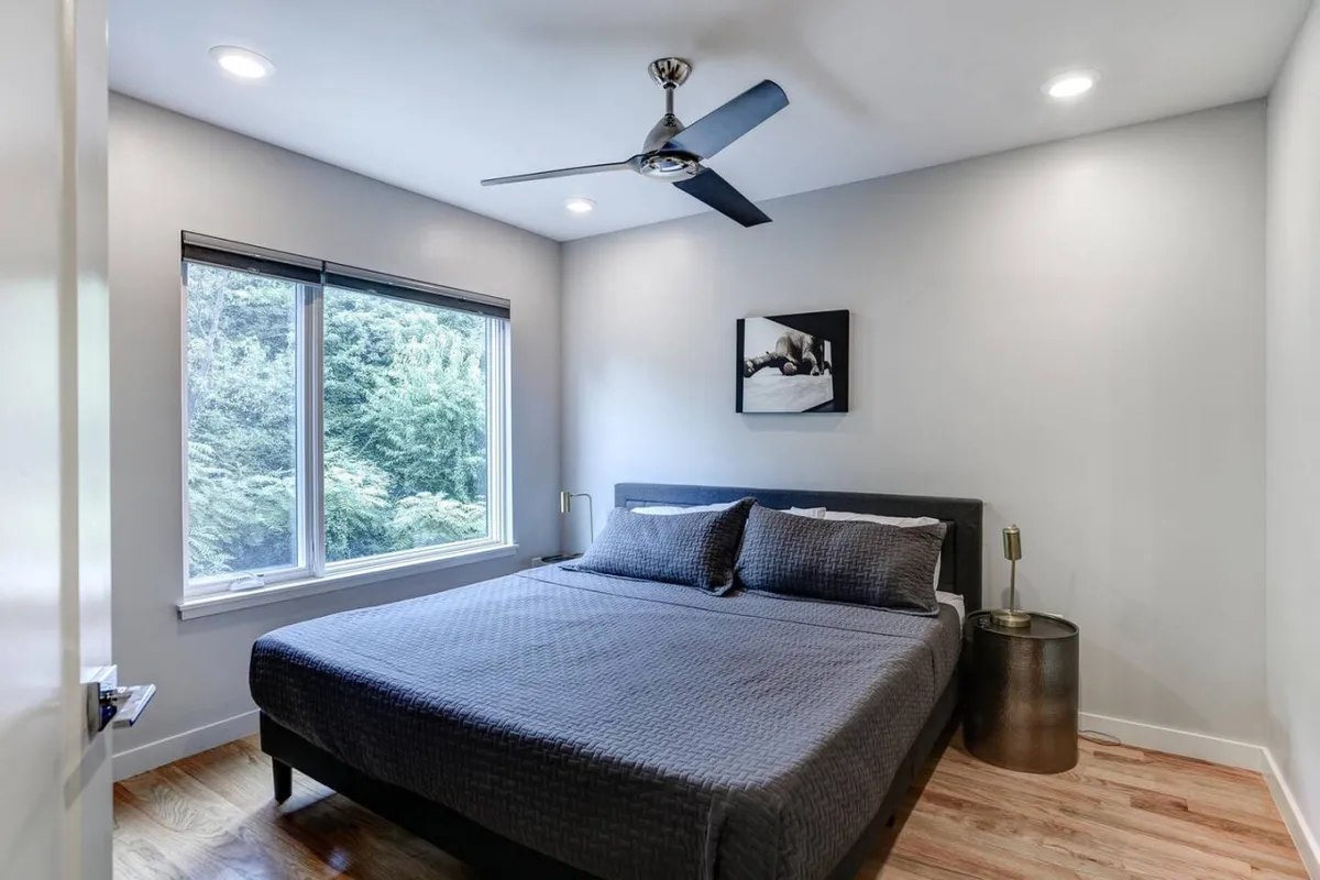 This image shows a serene bedroom with a large bed adorned with decorative pillows, artwork on the wall, and expansive windows offering a view of lush greenery. Natural light pours into the room, creating a calm and inviting atmosphere ideal for rest and relaxation.