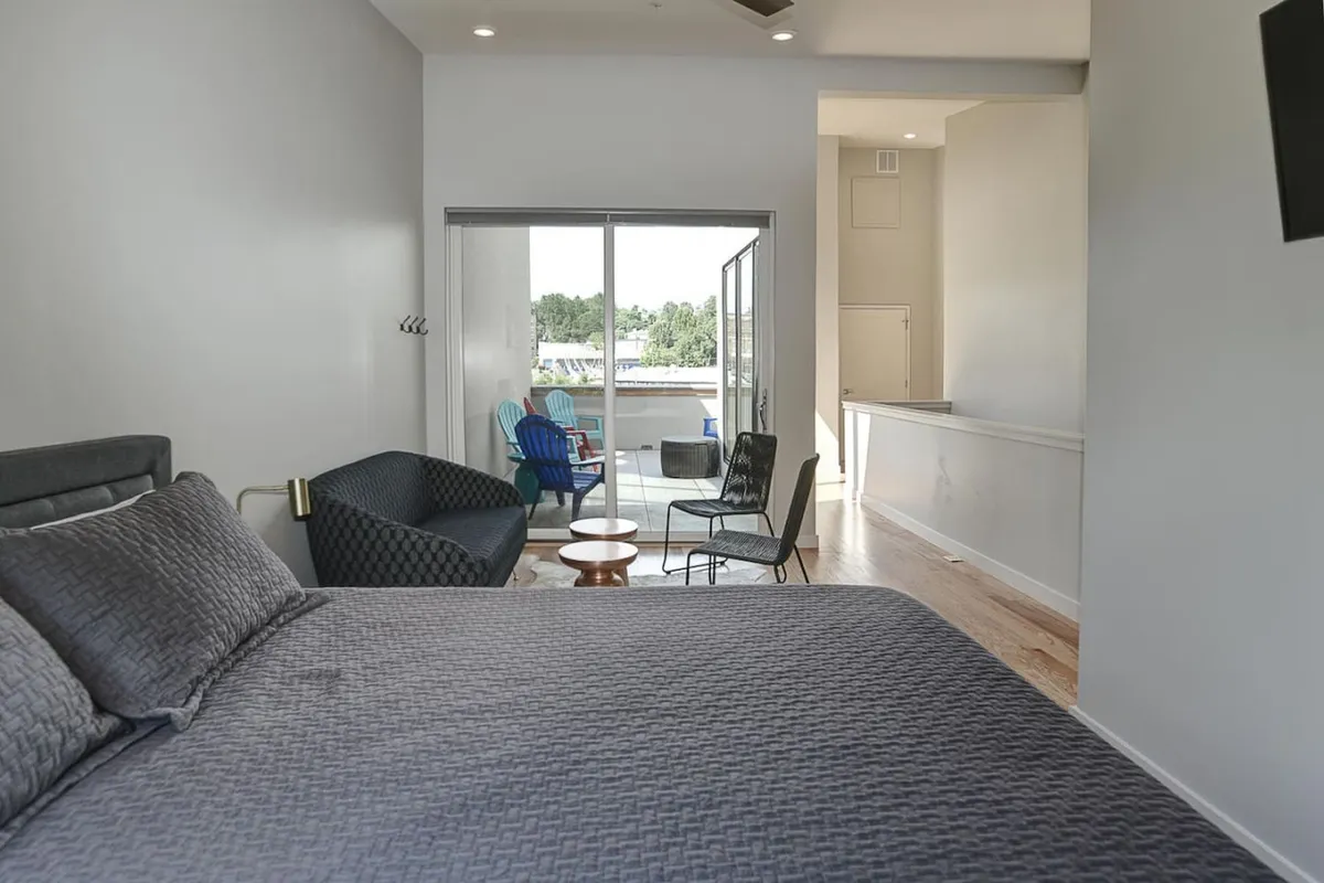 A cozy bedroom with a bed facing a small balcony furnished with chairs and a wooden table. The space is perfect for unwinding, reading, or enjoying morning coffee while taking in views from the balcony.