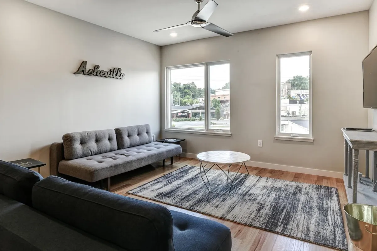 A contemporary lounge area featuring a plush sofa, a stylish coffee table, and decorative wall art. Expansive windows provide a view of the outside and bring in plenty of daylight, creating a welcoming spot for relaxation or lively conversation.